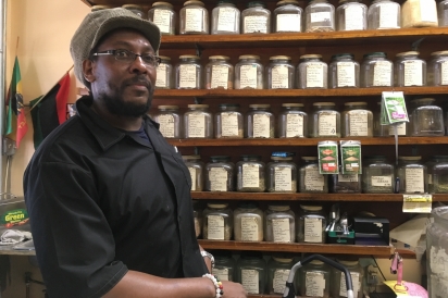 Co-owner of H.I.M Ital Oba Kenjah stands in front of a wall of herbs and spices 